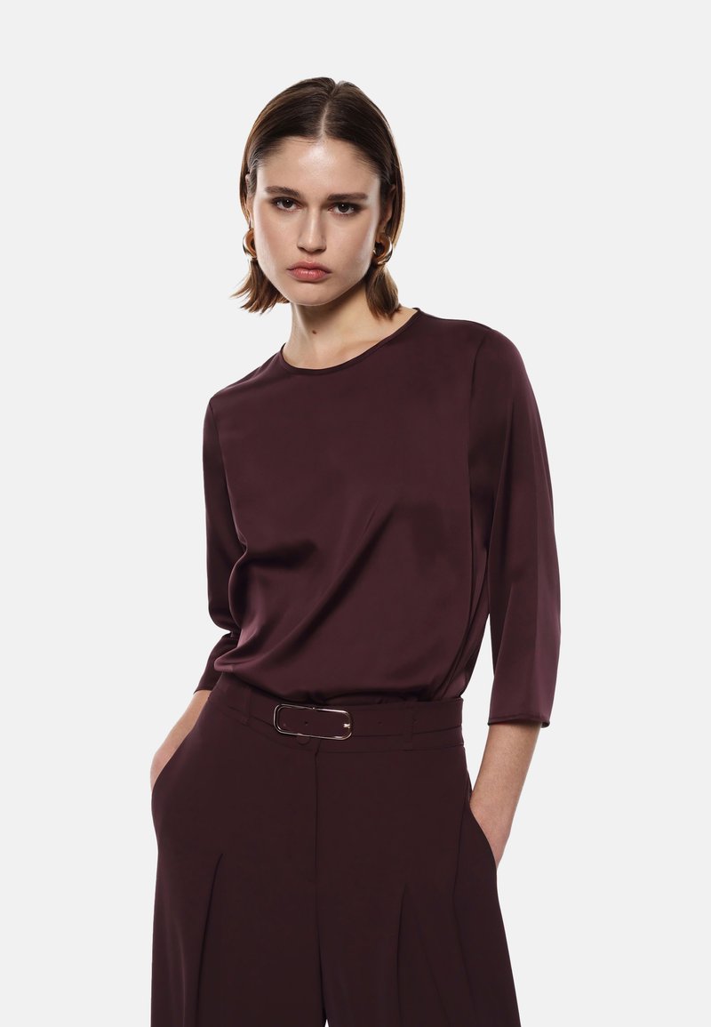Blusa bordeaux con maniche a tre quarti, texture liscia, scollatura rotonda e vestibilità morbida, abbinata a pantaloni a vita alta coordinati.
