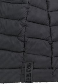 Schwarze Steppjacke aus Material mit einem Rautenmuster und quilted Abschnitten. Das Etikett trägt den Namen "Sublevel" und befindet sich unten links.