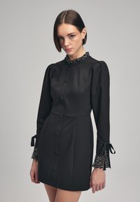 Robe noire à boutons avec une silhouette ajustée, des manches bouffantes et un col à pois avec une bordure en dentelle et des nouettes aux poignets.
