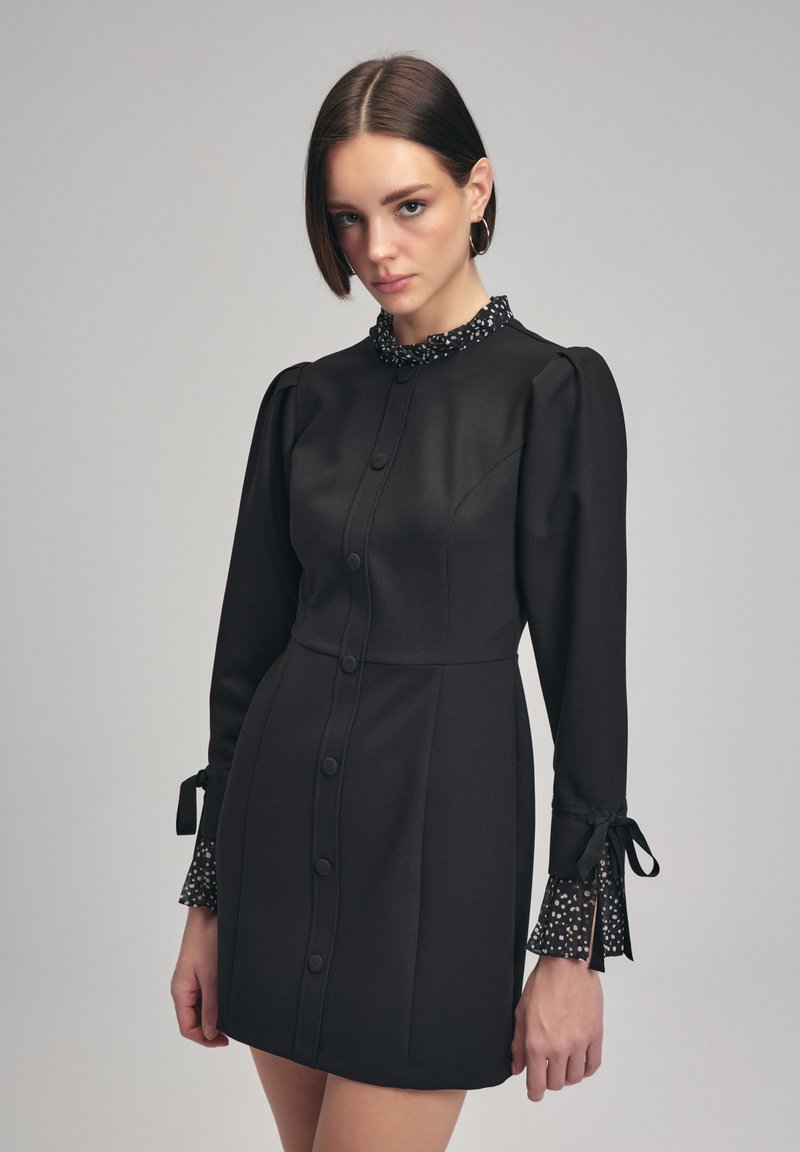 Robe noire à boutons avec une silhouette ajustée, des manches bouffantes et un col à pois avec une bordure en dentelle et des nouettes aux poignets.