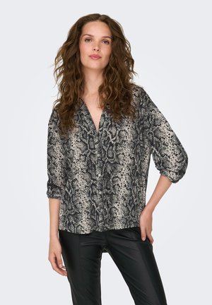 Femme aux cheveux bruns bouclés portant un chemisier ample boutonné à imprimé serpent et un pantalon en cuir noir, debout avec une main sur la hanche.