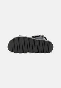 Black sandal with a textured rubber sole, featuring adjustable straps and metallic buckles, designed for comfort and stability.