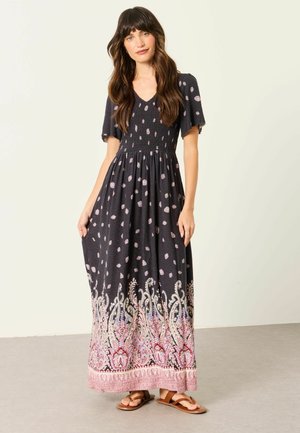 Femme aux longs cheveux bruns, vêtue d'une robe longue noire à motif floral rose et blanc et de sandales marron, debout devant un fond uni.