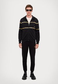 Black zip-up hoodie with gold stripes, paired with matching black sweatpants featuring logo detail. Model wears black sunglasses and shoes.