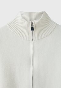 Pull en maille côtelée blanche avec un col haut et une fermeture éclair argentée partiellement fermée au niveau du cou sur un fond uni.