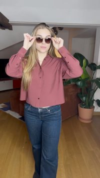 Camisa corta de color burdeos con botones y mangas largas, combinada con unos jeans acampanados de color azul oscuro. El modelo sostiene unas gafas de sol marrones, con plantas de interior en el fondo.