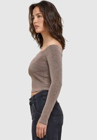 Off-the-shoulder metallic bronzen cropped top met lange mouwen, gecombineerd met donkere high-waisted jeans. Zachte textuur met een aansluitende snit.