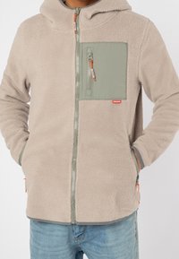 Personne portant une veste en polaire beige avec une poche poitrine zippée verte, les mains dans les poches latérales, associée à un jean bleu clair.