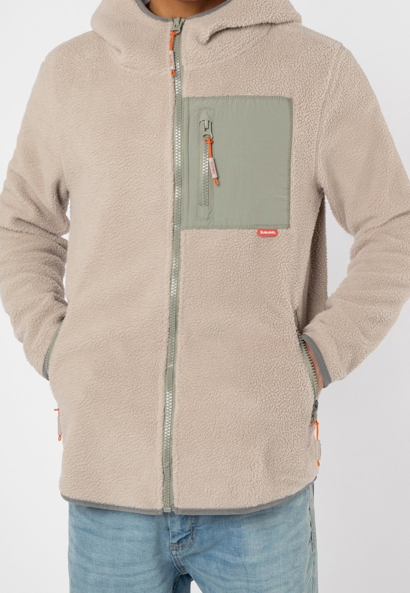 Personne portant une veste en polaire beige avec une poche poitrine zippée verte, les mains dans les poches latérales, associée à un jean bleu clair.