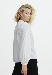 Weiße Bluse mit strukturiertem Punktmuster, hohem Kragen und langen Puffärmeln. Der Stoff wirkt leicht und leicht durchscheinend.