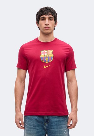 Ung mand med kort krøllet hår iført en rød FC Barcelona t-shirt og blå jeans står mod en ensfarvet lys baggrund.