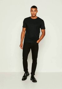 Mees seisab heleda tausta ees, seljas must lühikeste varrukatega särk, mustad püksid ja mustad nahksaapad, ühe käega taskus.