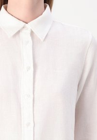 Camicia bianca in lino con colletto a punta, chiusura con bottoni e una texture sottile. Presenta un piccolo logo ricamato sul petto.
