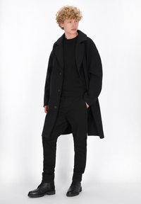 Jonge man met krullend haar, gekleed in een zwarte jas, zwarte broek en zwarte laarzen, poseert met één hand in zijn zak tegen een witte achtergrond.