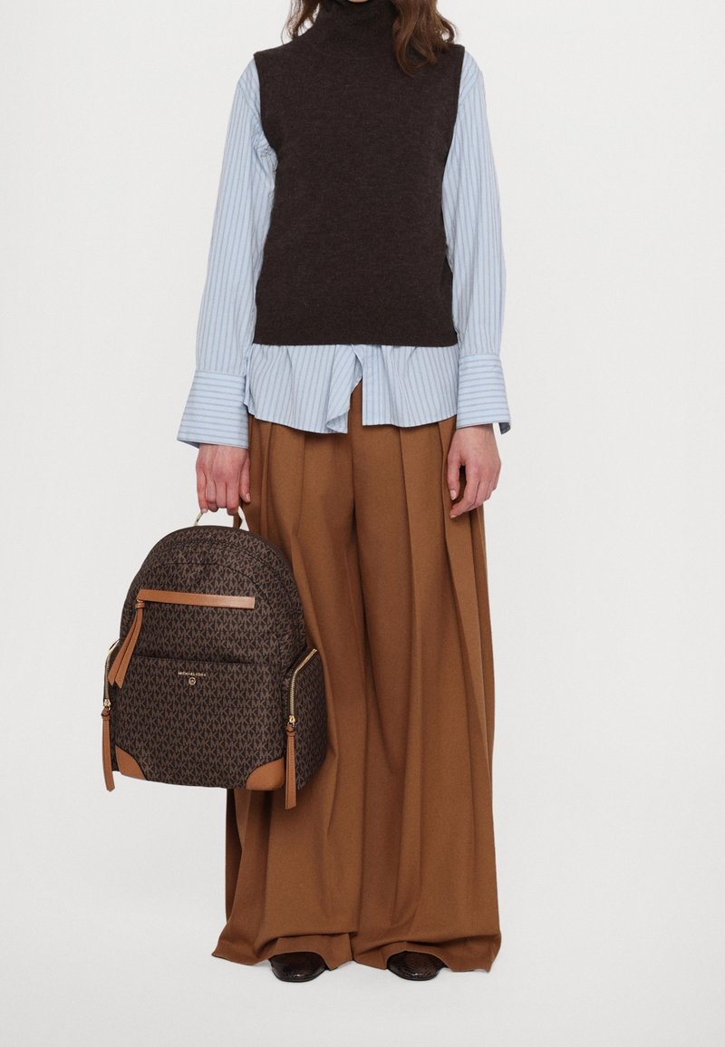 Personne portant une chemise rayée bleu clair, un pull sans manches marron foncé, un pantalon large marron, tenant un sac à dos marron à motifs.