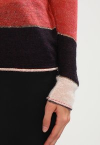 Gehaakte trui met een kleurblokontwerp in tinten van koraal, donkerpaars en room. Kenmerken ribgebreide manchetten en metalen draadaccenten langs de randen.