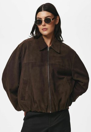 Femme portant une veste zippée en daim marron foncé et des lunettes de soleil rondes noires, debout les mains derrière le dos sur un fond blanc uni.