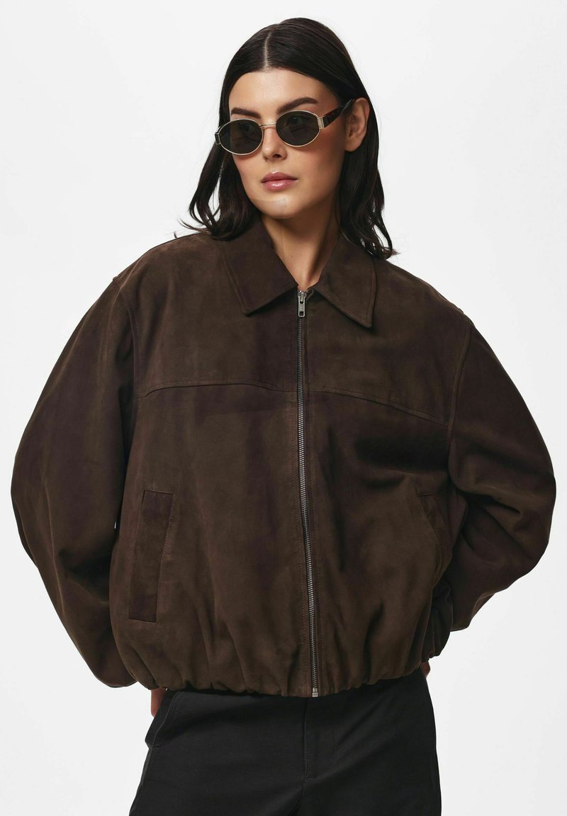 Femme portant une veste zippée en daim marron foncé et des lunettes de soleil rondes noires, debout les mains derrière le dos sur un fond blanc uni.