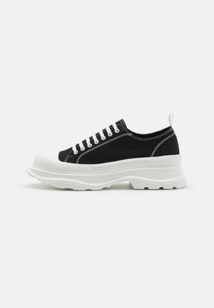 Pantofi sport negri din pânză, cu talpă de cauciuc albă, vârful rotund și șireturi albe. Talpă distinctiv texturată și detalii de cusătură pe lateral.