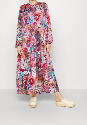 Femme portant une robe longue à manches longues avec des motifs floraux et de feuilles colorés, associée à des sabots en bois beige.