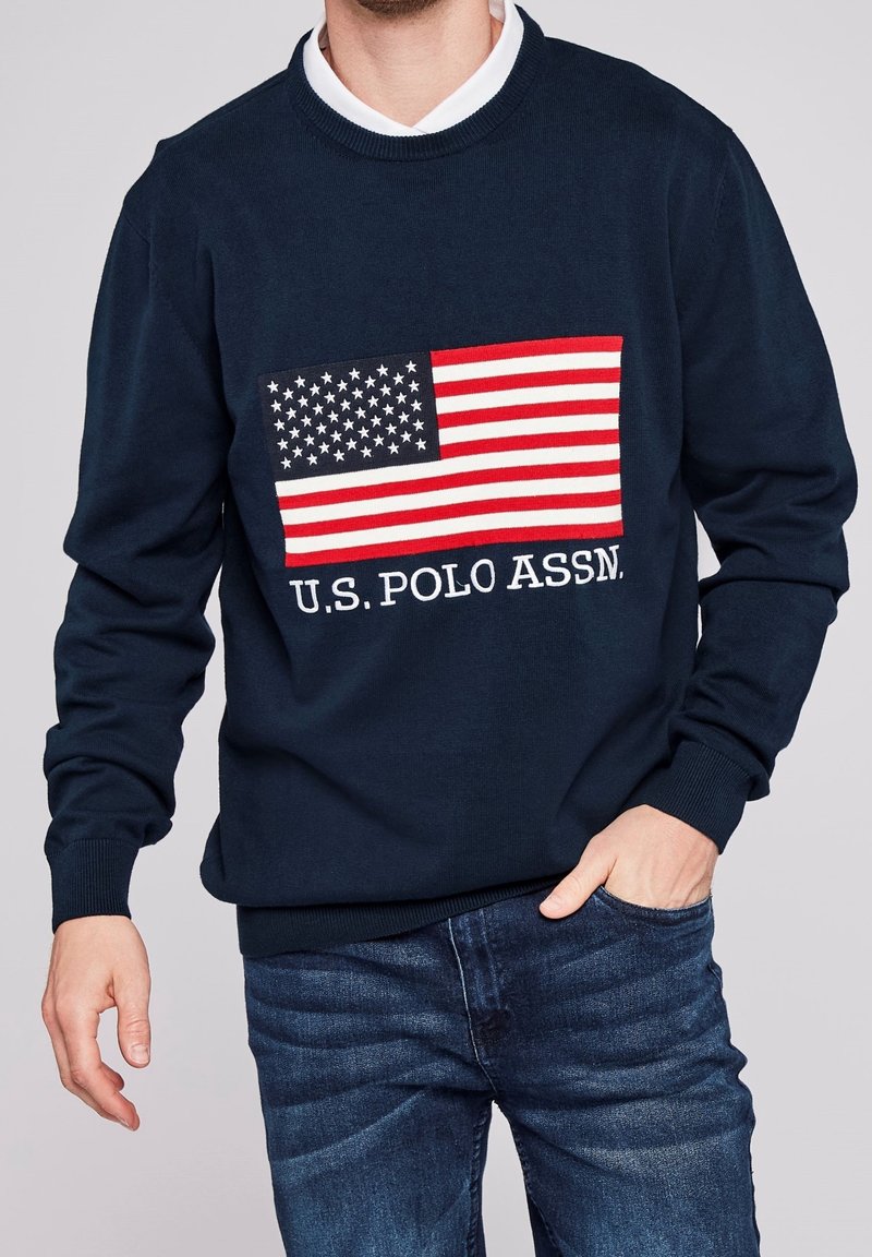 Jersey de punto azul marino con un gráfico de la bandera americana y el texto "U.S. POLO ASSN.", con puños y dobladillo acanalados, que complementa unos vaqueros de mezclilla.