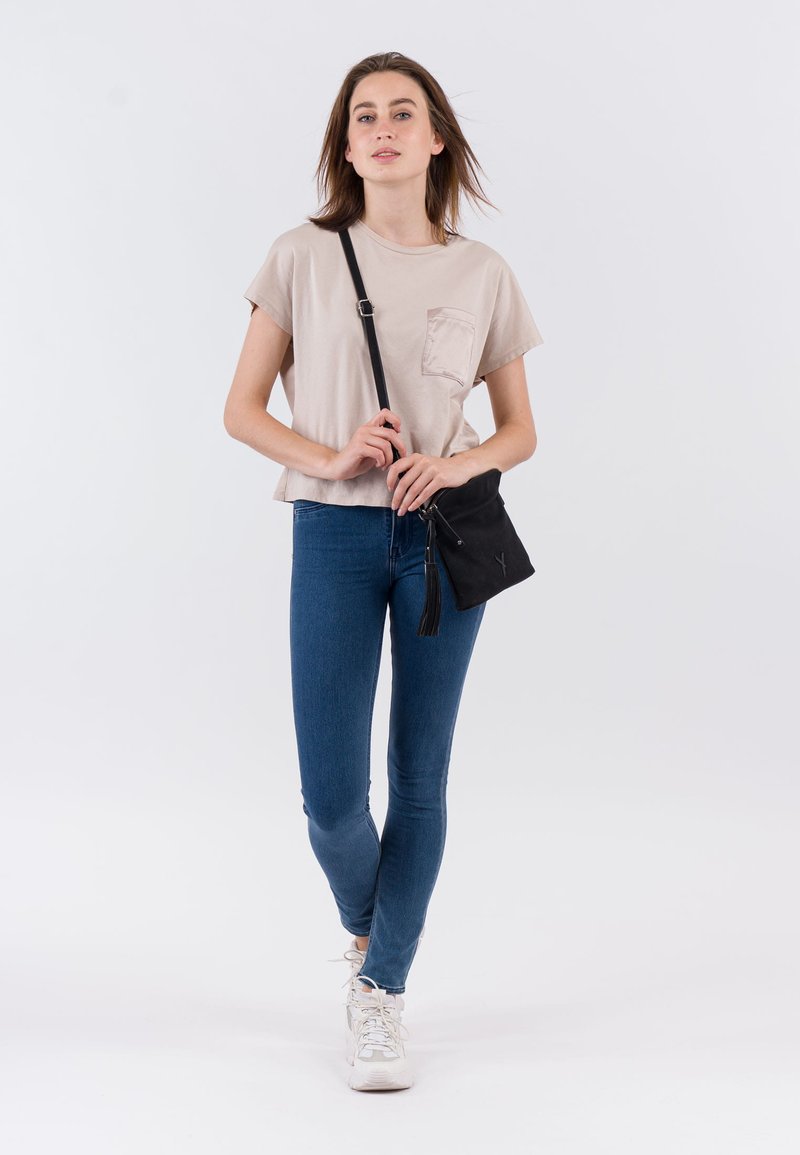 Moteris stovinti, vilkinti smėlio spalvos marškinėlį, mėlinais siaurais džinsais, baltomis sportbačiais ir laikanti juodą per petį nešiojamą rankinę su kutais.