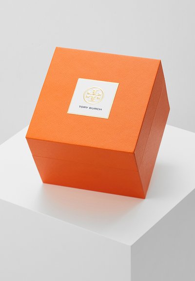 Scatola quadrata arancione con texture con logo Tory Burch bianco e oro sul coperchio, esposta su un piedistallo bianco.
