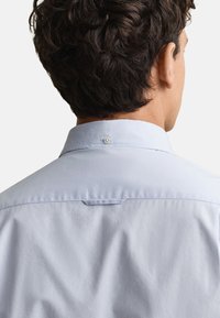 Homme portant une chemise bleu clair à col boutonné, vu de dos, avec des cheveux courts, bouclés et foncés sur un fond uni.