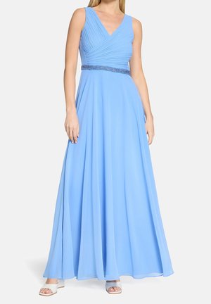 Femme portant une robe longue bleu clair sans manches avec un corsage plissé et une ceinture perlée, associée à des talons blancs ouverts à l'avant.