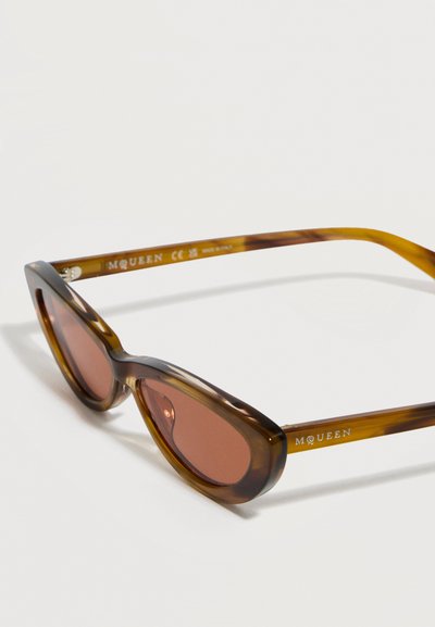 Καφέ γυαλιά ηλίου με σχέδιο γατίσιου ματιού και καφέ φακούς, με την επωνυμία «McQueen» στα πλαϊνά, τοποθετημένα πάνω σε λευκή επιφάνεια.