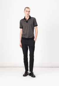 Mustard- ja beež houndstooth-mustriga polo särk musta krae ja varrukaotstega, koos kitsaste mustade pükste ja mustade kingadega.