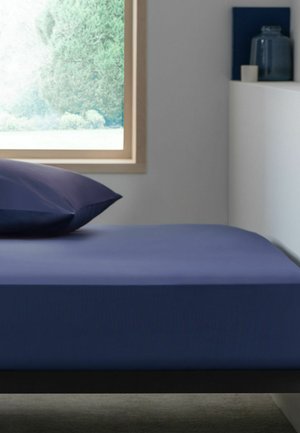 Lit simple avec une literie et un oreiller bleu marine à côté d'une fenêtre montrant un feuillage vert à l'extérieur dans une pièce simple et moderne.