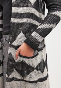 Dlouhý cardigan s černobílým geometrickým vzorem, pletenou texturou a kapsou na levé straně. Design s otevřeným předním dílem.