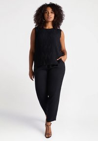 Zwarte mouwloze top met gestructureerde plooien, gecombineerd met zwarte broek. Het model draagt strappy zwarte en bruine geprinte hakken.