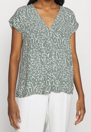 Femme portant un chemisier vert à manches courtes avec un motif abstrait blanc et un col en V, associé à un pantalon blanc, debout devant un fond uni.
