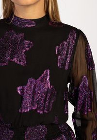 Zwarte jurk met paarse bloemenborduursels. Lange doorschijnende mouwen, hoge hals en textuurstof. Heeft een smocked taille voor een getailleerde vorm.