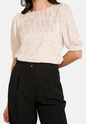 Femme portant un chemisier court texturé crème à manches bouffantes, rentré dans un pantalon noir plissé taille haute avec poches latérales.