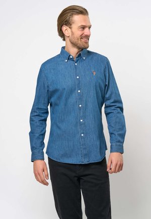 Mann smiler og ser mot høyre, iført en blå denimskjorte med knapper og svarte bukser mot en enkel, lys bakgrunn.