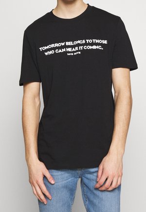 Henkilö, jolla on päällään musta T-paita, jossa valkoisella teksti "Tomorrow belongs to those who can hear it coming. David Bowie", ja siniset farkut.