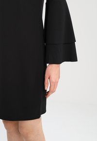 Robe noire avec manches cloche superposées. Le matériau semble lisse avec une coupe structurée. Main placée près de l'ourlet, montrant le détail des manches.