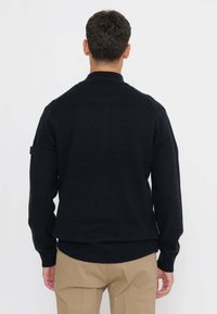 Uomo con maglione nero a maniche lunghe e pantaloni beige, visto di spalle su uno sfondo bianco semplice.