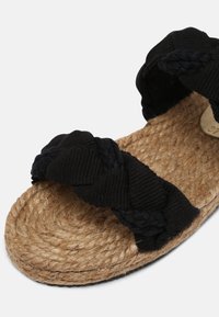 Sandales en tissu noir tressé avec une semelle en corde texturée. La partie supérieure présente une sangle en tissu doux pour le confort, reposant sur une base en jute naturel.