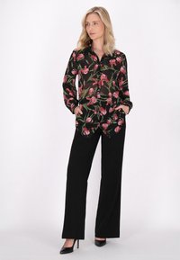 Bluză neagră cu un design floral roz și frunze verzi, cu nasturi la partea din față, mâneci lungi și o margine care coboară pe laterale. Pantaloni negri, largi.