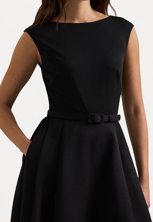 Frau trägt ärmelloses schwarzes Kleid mit Boots-Ausschnitt und passendem Gürtel, Hand in der Tasche, vor einfachem hellem Hintergrund.