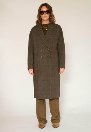 Manteau croisé à carreaux marron, coupe longue, larges revers et poches latérales, porté sur une tenue vert olive avec des chaussures marron.