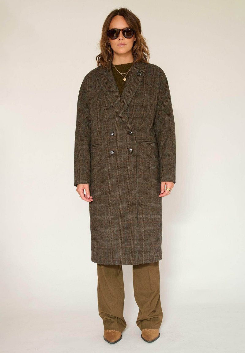 Manteau croisé à carreaux marron, coupe longue, larges revers et poches latérales, porté sur une tenue vert olive avec des chaussures marron.