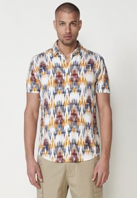 Chemise à manches courtes avec un motif géométrique multicolore en orange, bleu marine et blanc ; dotée d'un col boutonné et de boutons en bois.