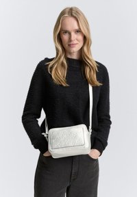 Borsa a tracolla bianca in pelle con testo impresso, forma rettangolare, tracolla regolabile e texture liscia. Modello che indossa un maglione nero e jeans.