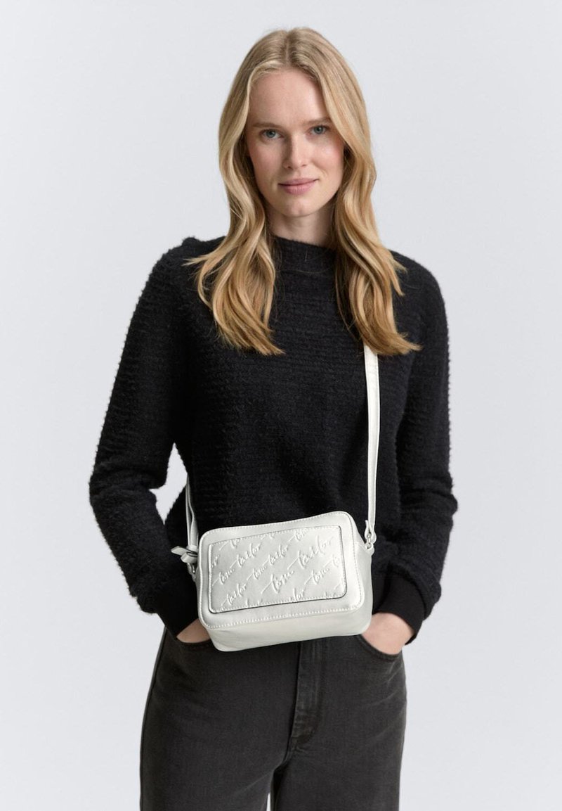 Borsa a tracolla bianca in pelle con testo impresso, forma rettangolare, tracolla regolabile e texture liscia. Modello che indossa un maglione nero e jeans.