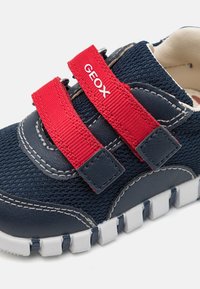 Scarpa sportiva navy con strap in velcro rossi, tomaia in tessuto testurizzato, suola in gomma bianca e dettagli di cucitura a contrasto. Marchio Geox visibile.
